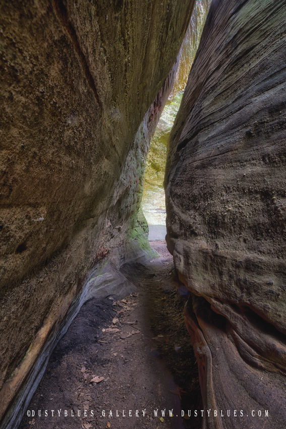 A photo of a passageway through two rocks that is not very wide leading to a sunlit rock face beyond